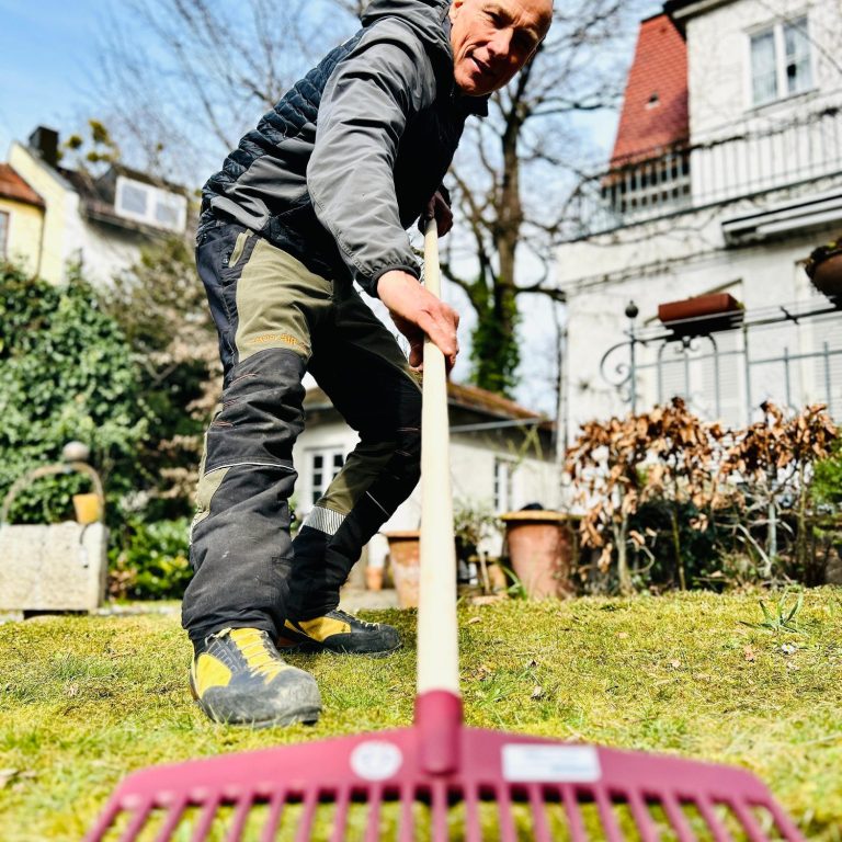 Patrick Weihe mit Rechen auf dem Rasen in einem Garten, Sonnenschein.