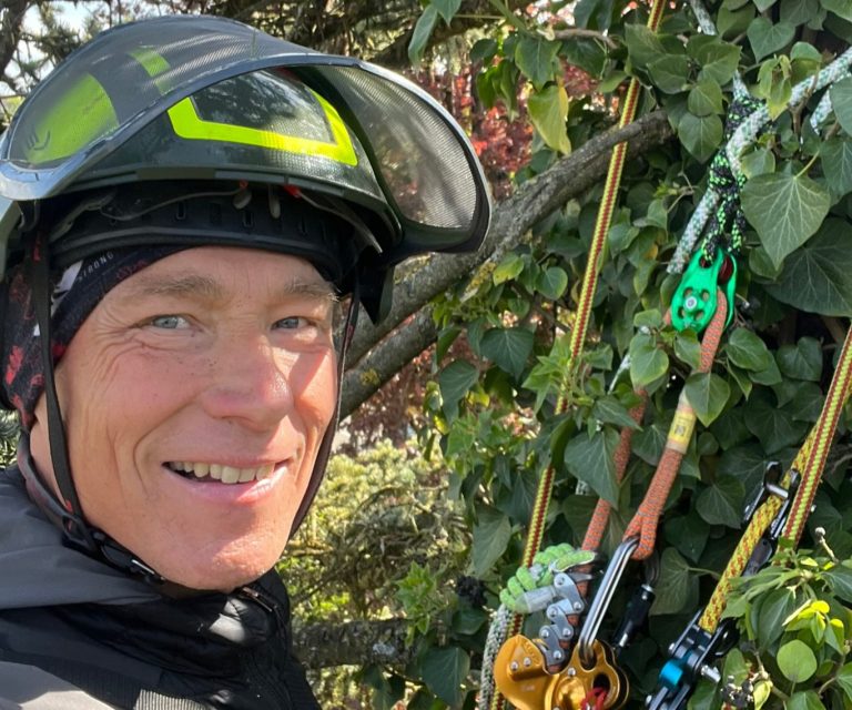 Lächelnder Patrick Weihe mit Schutzhelm, umgeben von grünen Pflanzen und Kletterausrüstung.
